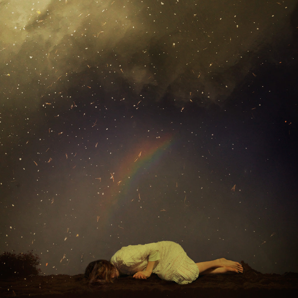 A young woman in a white dress is curled up on the ground beneath a starry night sky.