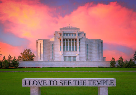 A large white temple building stands on a green lawn under a dramatic pink, orange, and purple sunset sky. In the foreground, a stone sign reads, 'I LOVE TO SEE THE TEMPLE'.