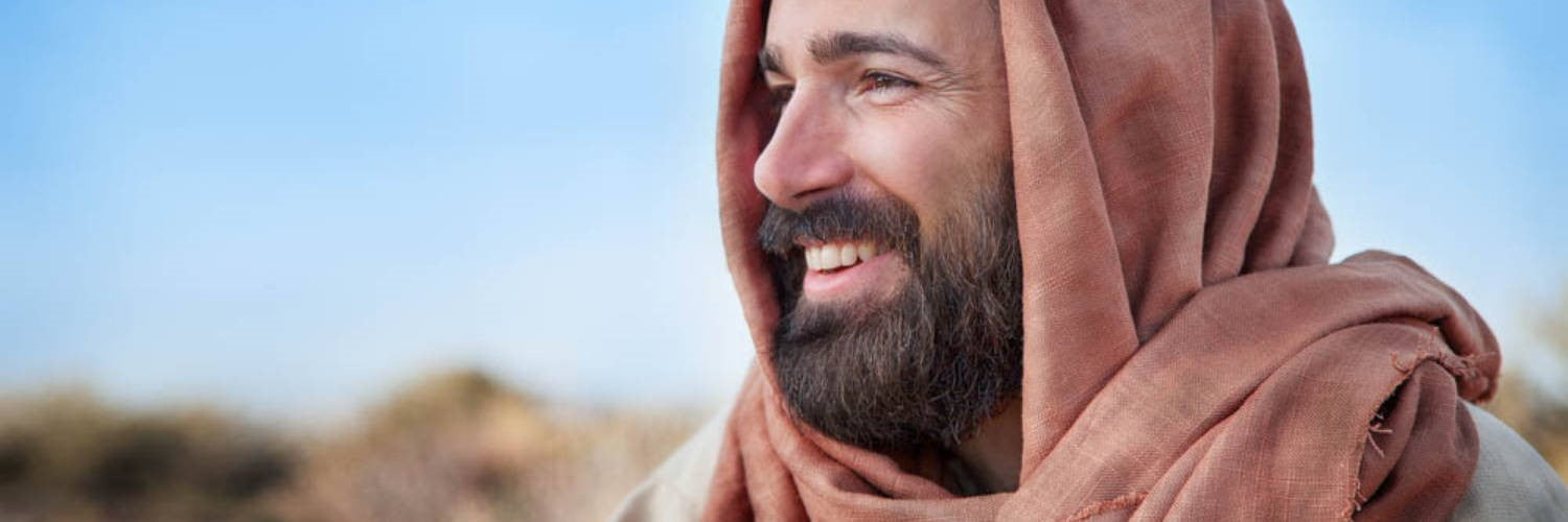 A smiling man with a beard wearing a traditional brown head covering and robe, looking off to the side against a soft outdoor background.