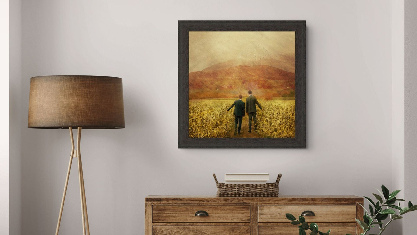 Framed artwork of two people walking in a field above a wooden dresser with a lamp and plant.