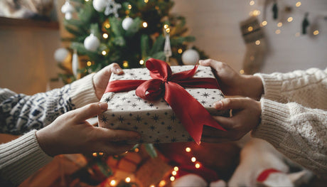 Two people exchanging a wrapped Christmas present with a red bow in front of a decorated Christmas tree.