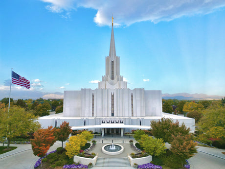 The Jordan River Utah Temple surrounded by fall trees and a blue sky.