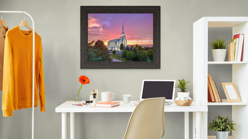 Home office with a framed picture of a sunset over a building, a desk with a laptop, and a sweater on a rack.