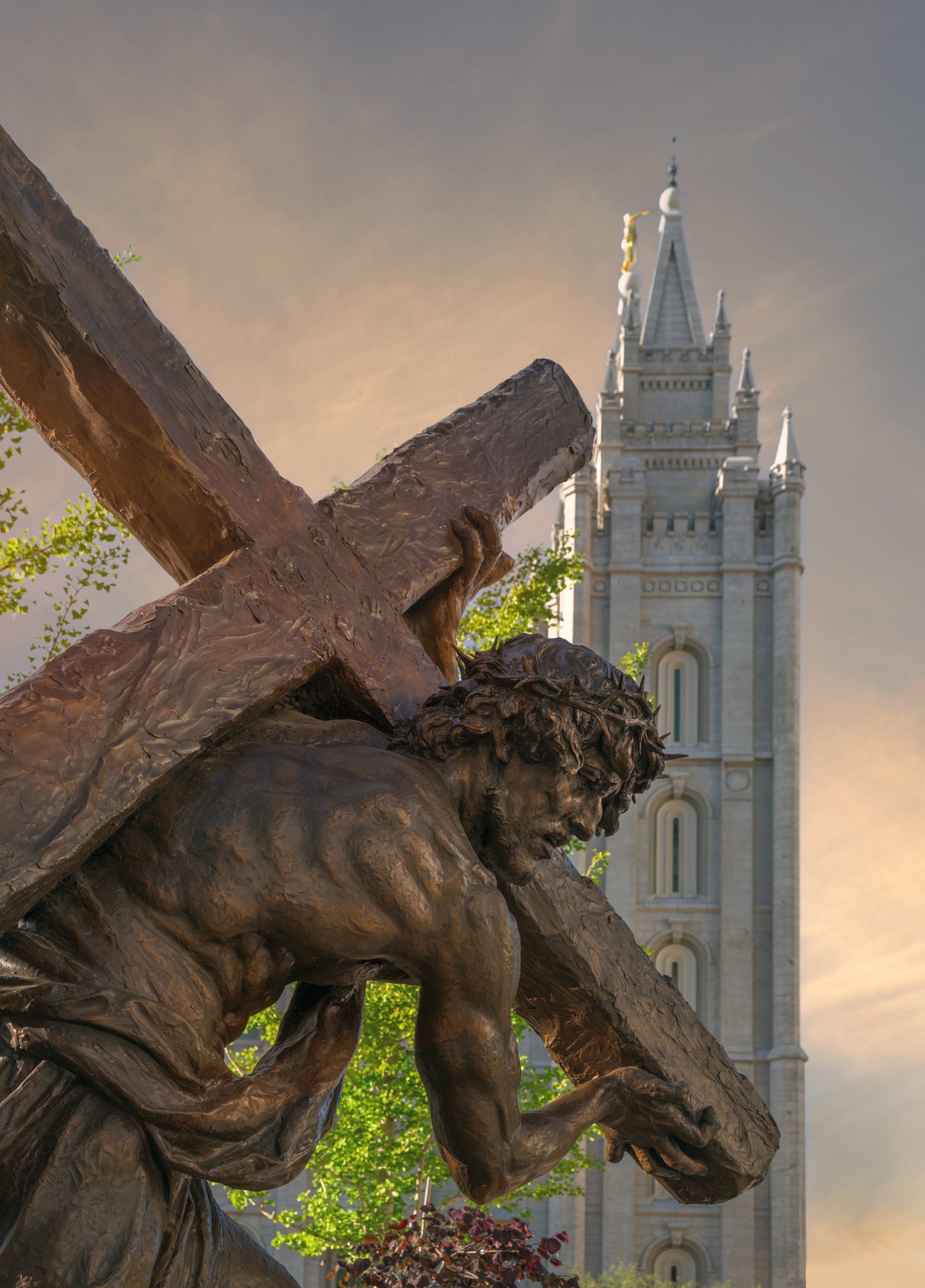 A bronze statue of Christ carrying the cross, shown from a low angle with the Salt Lake Temple spire visible in the background against a warm sunset sky