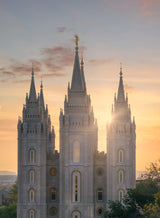 Photograph of the Salt Lake Temple's spires at sunrise, sunlight beaming through between the spires with the golden Angel Moroni statue silhouetted at the top, the carved 'Holiness to the Lord' inscription visible on the center spire.