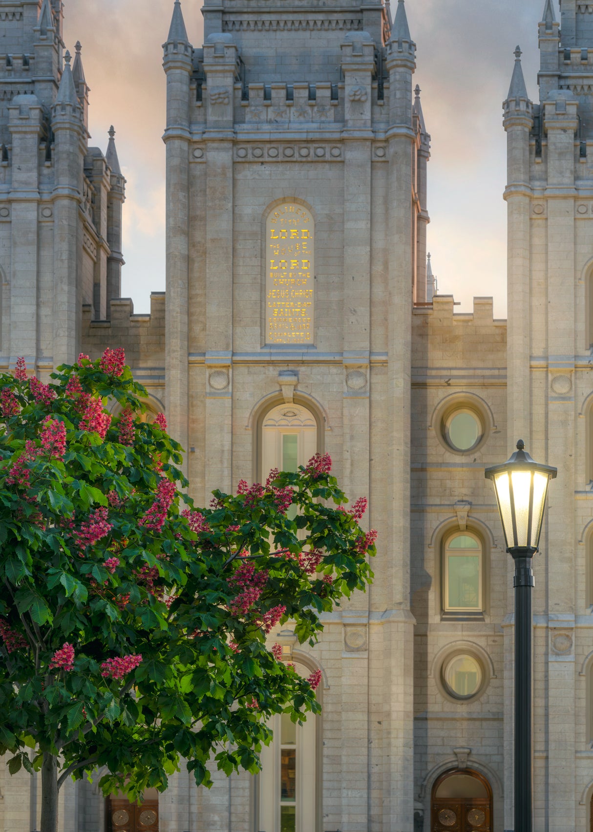 Photograph of the Salt Lake Temple's stone facade with the carved inscription 'Holiness to the Lord, the House of the Lord' near the top, a flowering pink tree and a lamppost in the foreground.