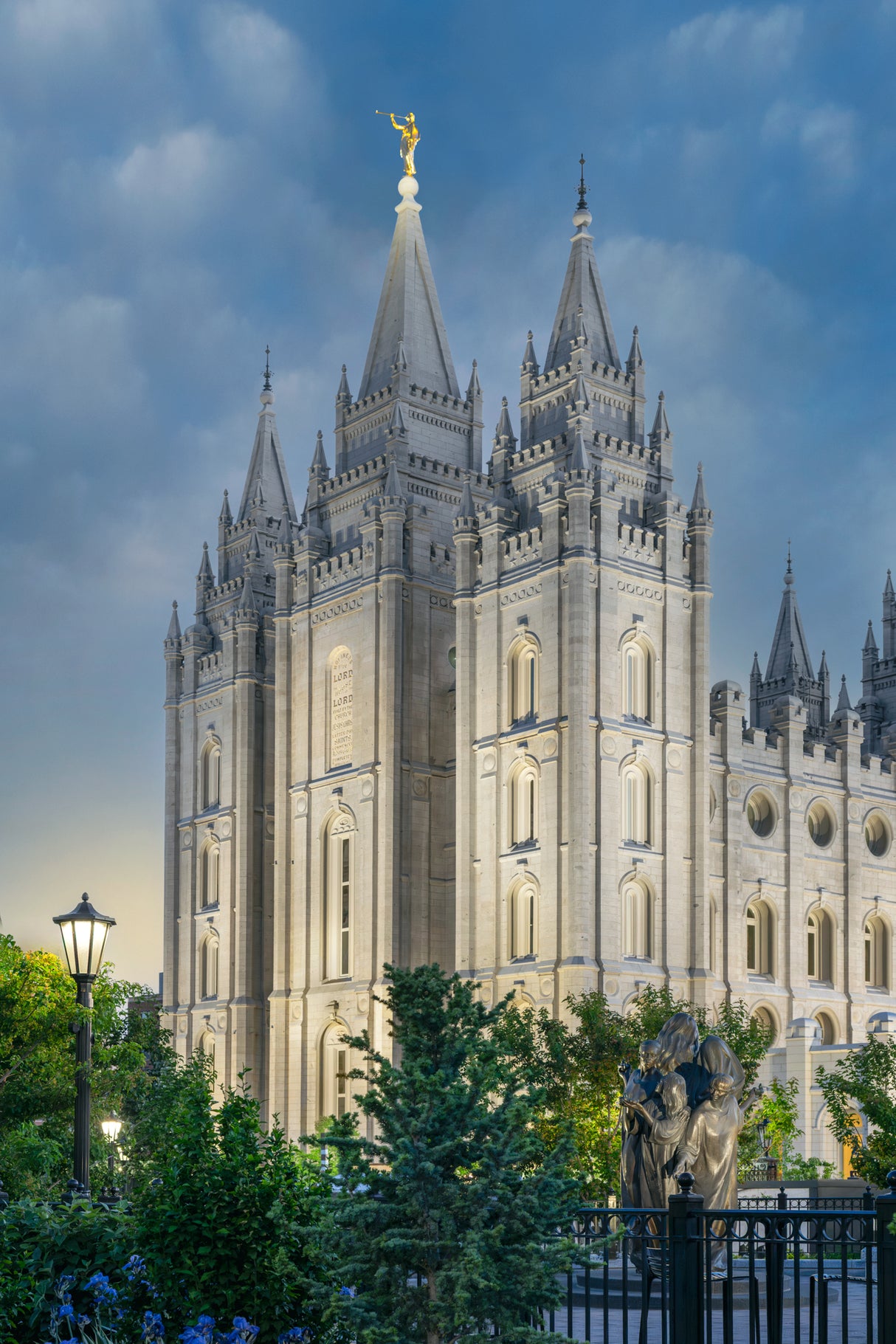 Photograph of the Salt Lake Temple lit up at dusk against a blue sky, with a lamppost and trees in the foreground and an iron fence bordering the grounds.
