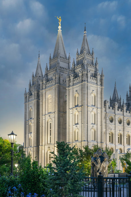 Photograph of the Salt Lake Temple lit up at dusk against a blue sky, with a lamppost and trees in the foreground and an iron fence bordering the grounds.