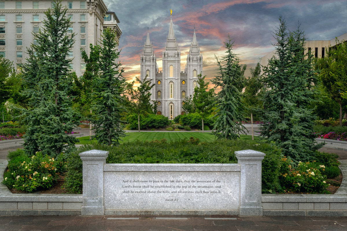 Photograph of the Salt Lake Temple at sunset, framed by evergreen trees and a green lawn with a pink and orange cloudy sky behind its spires. A granite wall in the foreground is inscribed with Isaiah 2:2, 'the mountain of the Lord's house shall be established in the top of the mountains,' with the Joseph Smith Memorial Building visible at the left.