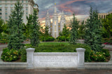 Photograph of the Salt Lake Temple at sunset, framed by evergreen trees and a green lawn with a pink and orange cloudy sky behind its spires. A granite wall in the foreground is inscribed with Isaiah 2:2, 'the mountain of the Lord's house shall be established in the top of the mountains,' with the Joseph Smith Memorial Building visible at the left.