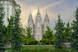 Photograph of the Salt Lake Temple with a faint rainbow arcing across a cloudy sky behind its three spires, the golden Angel Moroni glowing at the top. Evergreen trees and a manicured lawn frame the temple, with the Joseph Smith Memorial Building rising at the left edge