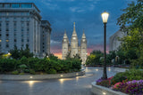 Photograph of the Salt Lake Temple at twilight under a deep blue sky with a band of orange and pink on the horizon. A curved stone path lined with flowerbeds and a glowing lamppost leads toward the temple, with the Joseph Smith Memorial Building at the left and the Church Office Building at the right.