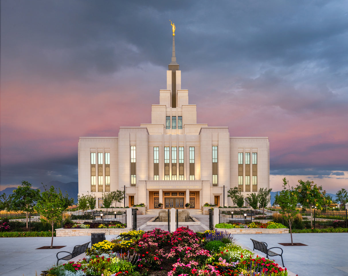 A photograph of the Saratoga Springs Utah Temple glowing at sunset, with colorful flower gardens lining the walkway to the entrance beneath a pink and orange sky.