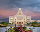 A photograph of the Saratoga Springs Utah Temple glowing at sunset, with colorful flower gardens lining the walkway to the entrance beneath a pink and orange sky.