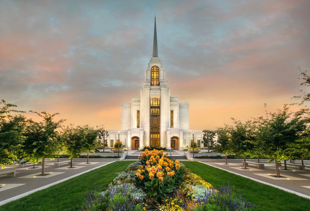 Syracuse Utah Temple glowing warmly at sunset, with a manicured garden path lined with orange and purple flowers leading up to the entrance beneath a pastel sky.