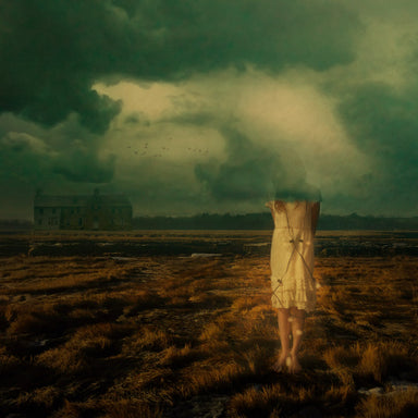 A young woman stands in a field. Her face is blocked out by low-hanging clouds. 
