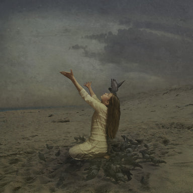 A girl sits on a stormy beach with her face upturned and her arms upstretched toward Heaven. She is surrounded by black birds