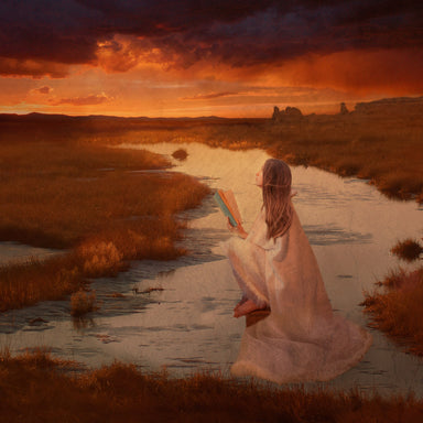 A young woman sits on a stone in a riverbed, holding an open book and looking up at the evening sky.