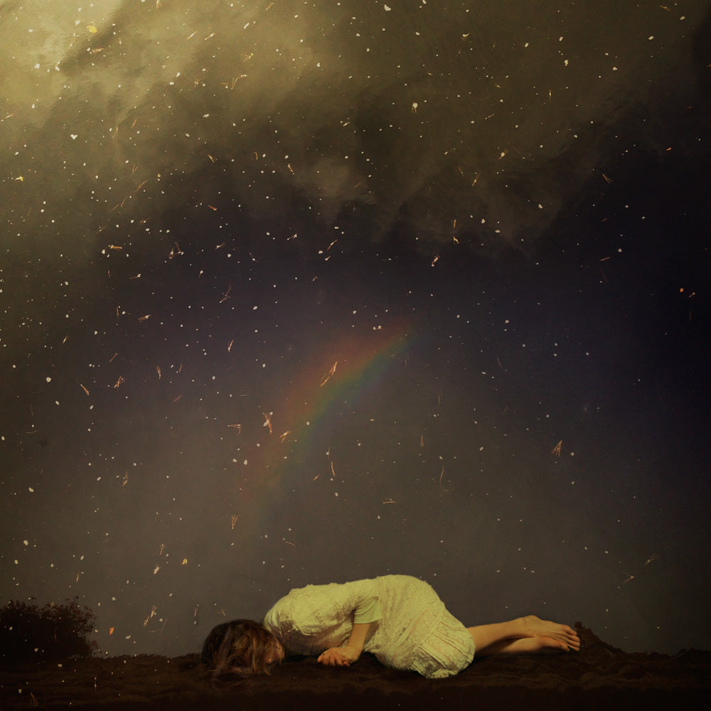 A young woman in a white dress is curled up on the ground beneath a starry night sky. 
