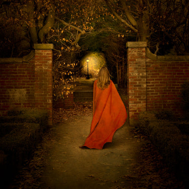 A young woman in a red cloak holds a light as she ventures down a garden path.
