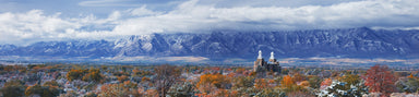 A panoramic landscape of the Logan Utah Temple surrounded by fall trees with mountains in the background.